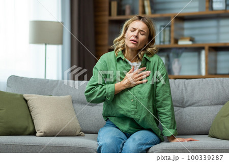 Heart attack, panic attack,. An elderly woman sits on the sofa at home and holds her hand to her chest. It is difficult to breathe, feels severe pain. 100339287