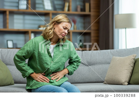 Unhealthy senior woman sitting on sofa at home. holding her stomach, grimacing in pain. She experiences severe abdominal pain, eating disorder, diarrhea, poisoning, menstruation, menopause. 100339288