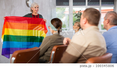 Focused adult students sitting at lesson with classmates, listening mature teacher talking about LGBT community and showing rainbow flag Focused adult students sitting at lesson with classmates, listening mature teacher talking about LGBT community and showing rainbow flag 100345806