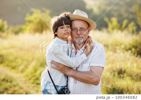 Photo of little boy and his grandfather standing and hugging in the field. Boy and grandfather spending time together at summer. Brunette boy and man wearing striped shirts. 100346082