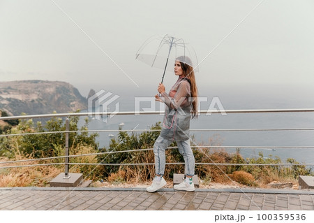 Woman rain umbrella. Happy woman portrait wearing a raincoat with transparent umbrella outdoors on rainy day in park near sea. Girl on the nature on rainy overcast day. 100359536