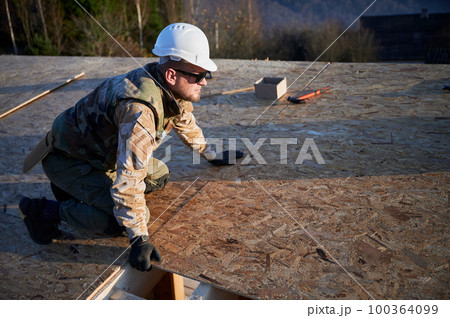Carpenter mounting wooden OSB board on rooftop of future cottage. Man worker building wooden frame house. Carpentry and construction concept. Carpenter mounting wooden OSB board on rooftop of future cottage. Man worker building wooden frame house. Carpentry and construction concept. 100364099