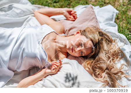 woman sleeps on a white bed in the fresh spring grass in the garden. Dressed in a blue nightgown. 100370919