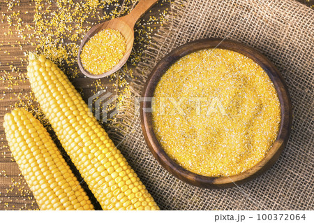 Corn grits in a wooden bowl on the background of fresh corn on the cob on a burlap. 100372064