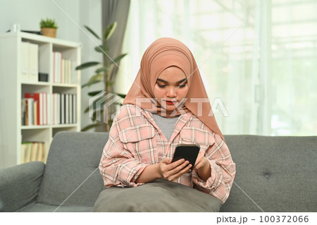 Charming young Muslim woman in hijab typing massage on her mobile phone while sitting on couch at home 100372066