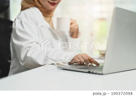 Cropped shot of smiling young muslim woman drinking coffee and checking email on laptop at her office desk 100372098