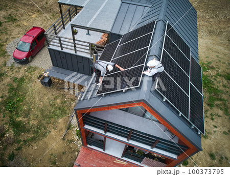 Engineers building photovoltaic solar module station on roof of house. Men electricians in helmets installing solar panel system outdoors. Concept of alternative and renewable energy. Aerial view. 100373795