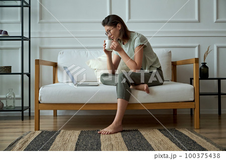 Young woman using laptop, sitting at home on sofa and drinking coffee, remote work, work at home Young woman using laptop, sitting at home on sofa and drinking coffee, remote work, work at home 100375438