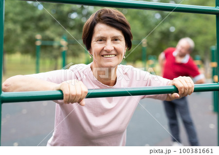 Aged woman doing exercises on sports bars in open-air sports area 100381661