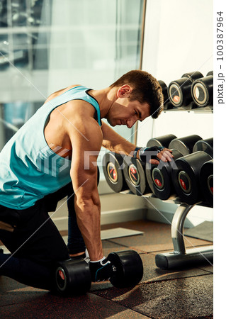Man doing exercise with dumbbell leaning on sports bench in the gym. Photo of a sexy muscular man in sportswear and good physique on grey background. Strength and motivation, sport, fitness goal Man doing exercise with dumbbell leaning on sports bench in the gym. Photo of a sexy muscular man in sportswear and good physique on grey background. Strength and motivation, sport, fitness goal 100387964