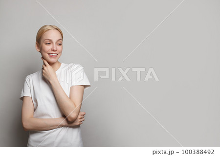 Attractive blonde female model in white t-shirt looking aside and smiling against grey studio wall  100388492