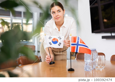 Positive young woman putting flag of South Korea on the table with flag of Norway in conference room 100392474