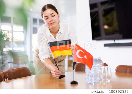 Positive young woman putting flag of Germany on the table with flag of Turkey in conference room Positive young woman putting flag of Germany on the table with flag of Turkey in conference room 100392525