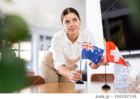 Young female assistant putting up swiss and new zealand flags on table for international negotiations Young female assistant putting up swiss and new zealand flags on table for international negotiations 100392536