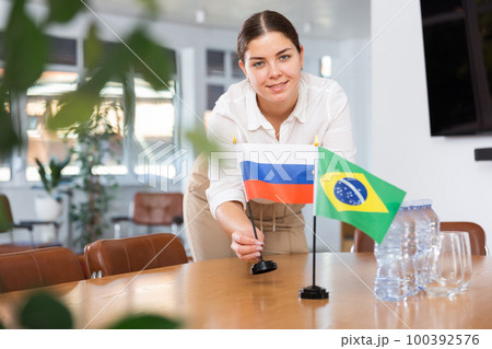 Positive young woman putting flag of Russia on the table with flag of Brazil in conference room 100392576