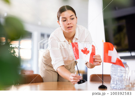 Positive young woman putting flag of Canada on the table with flag of Switzerland in conference room Positive young woman putting flag of Canada on the table with flag of Switzerland in conference room 100392583