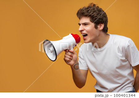 Young man shouting into megaphone making announcement against yellow background 100401344