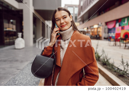 Cute brunette woman in brown coat talking on the phone in the city 100402419