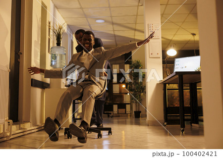 Cheeful employees having fun in startup office during work break, doing sports race competition with desk chair. Happy businessmen enjoying free time, acting playful late at night. Business concept Cheeful employees having fun in startup office during work break, doing sports race competition with desk chair. Happy businessmen enjoying free time, acting playful late at night. Business concept 100402421