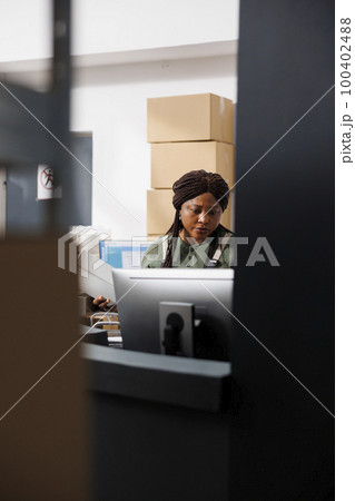 African american worker analyzing goods quality control report on computer, working at merchandise inventory in warehouse. Storage room employee preparing customers orders for delivery 100402488