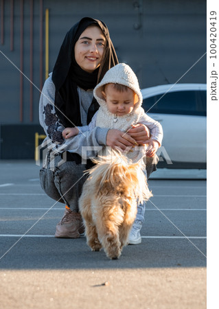 Woman in hijab enjoys walking with excited toddler and dog 100420419