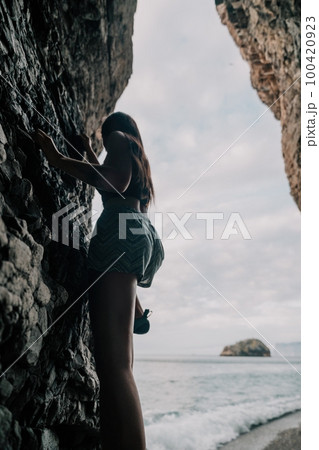 Sports Woman Climbing The Rock. Young woman With slim fit body climbing in volcanic basalt cave with beautiful sea view. The athlete girl trains in nature. Woman overcomes difficult climbing route. 100420923