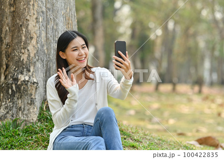 Pretty young asian woman making video call with her friends on mobile phone while sitting under a tree in public park Pretty young asian woman making video call with her friends on mobile phone while sitting under a tree in public park 100422835