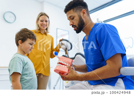 Young multiracial dentist showing little boy how to clean teeth, concept of children oral hygiene,prevention and dental health. Young multiracial dentist showing little boy how to clean teeth, concept of children oral hygiene,prevention and dental health. 100424948