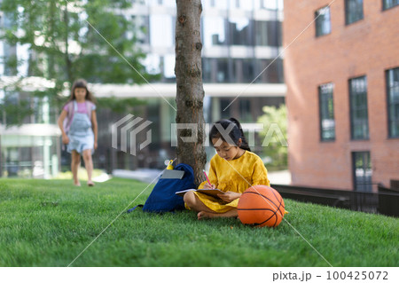 Little asian girl sitting in public park and writing notes, her friend standing in background. Summer time. 100425072