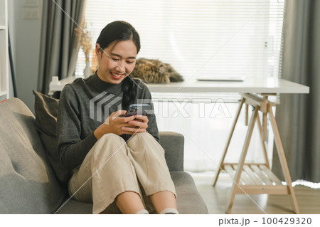 Charming Asian woman is using her smartphone while relaxing on the sofa at home. 100429320