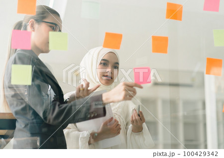 During a creative brainstorming process, business leaders are gathered in a boardroom, utilizing adhesive notes to exchange and enhance their ideas. 100429342