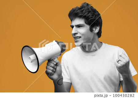 Young man shouting into megaphone making announcement against yellow background 100430282