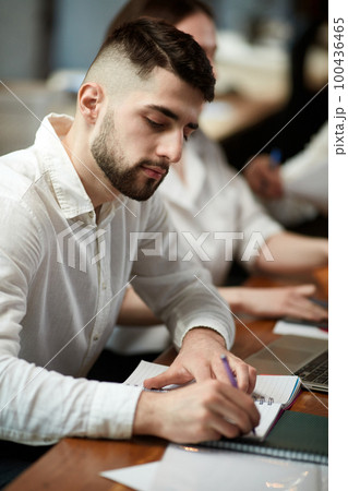 Young man, employee in formal clothes sitting at business meeting in office, making notes. Working on projects Young man, employee in formal clothes sitting at business meeting in office, making notes. Working on projects 100436465