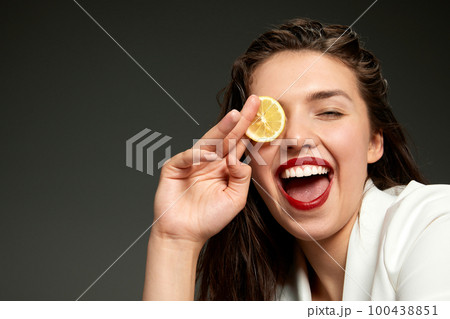 Emotional fashionable stylish beautiful woman wearing white jacket tasting slice of orange over gray background. Concept of healthy lifestyle, vitamins, dieting, fashion, beauty Emotional fashionable stylish beautiful woman wearing white jacket tasting slice of orange over gray background. Concept of healthy lifestyle, vitamins, dieting, fashion, beauty 100438851
