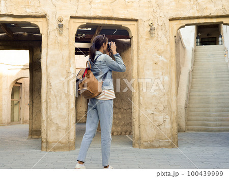 A young woman traveler with a backpack on her back walks and photographs the old narrow streets of Dubai Deira and Creek. Travel and sightseeing concept 100439999