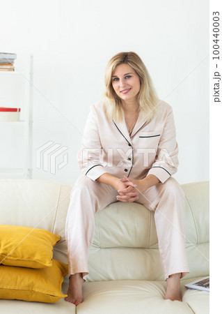 Smiling blonde woman sitting on comfortable coach in living room in pajama - homewear and free time holidays at home concept 100440003