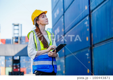 Female industrial engineer in uniform wearing safety hard hat using tablet checking containers. 100443871