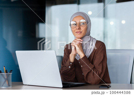 Portrait of successful and confident Muslim woman in hijab. Director, owner, business woman sitting in the office at a desk with a laptop. Portrait of successful and confident Muslim woman in hijab. Director, owner, business woman sitting in the office at a desk with a laptop. 100444386