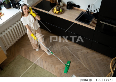 Happy beautiful young woman, housewife in rubber gloves, and white headphones, singing merrily while cleaning the kitchen floor Happy beautiful young woman, housewife in rubber gloves, and white headphones, singing merrily while cleaning the kitchen floor 100445571
