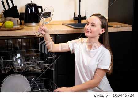 Beautiful young woman takes wine glasses out of the dishwasher and smiles while washing dishes in the kitchen Beautiful young woman takes wine glasses out of the dishwasher and smiles while washing dishes in the kitchen 100445724