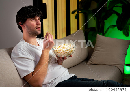 Young man sitting on the couch watching an interesting movie on the TV screen, eating popcorn Young man sitting on the couch watching an interesting movie on the TV screen, eating popcorn 100445971