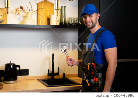Professional plumber fixing water tap in kitchen. Young professional plumber in grey uniform working on the kitchen 100445996