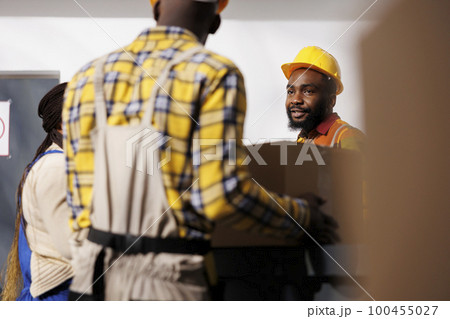 Warehouse manager looking at colleague bringing parcel to reception desk in storage room. African american storehouse employee wearing protective hard hat receiving box from package handler 100455027