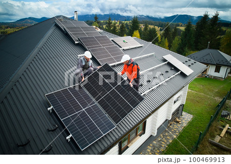 Engineers building photovoltaic solar module station on roof of house. Men electricians in helmets installing solar panel system outdoors. Concept of alternative and renewable energy. Aerial view. 100469913