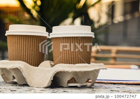 Two paper cups with lid for tea to go. Coffee take away on the table. Take-out coffees with brown paper cup holder. Brown safety cardboard collars. Cardboard tray Two paper cups with lid for tea to go. Coffee take away on the table. Take-out coffees with brown paper cup holder. Brown safety cardboard collars. Cardboard tray 100472747