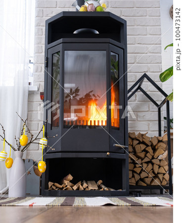 Easter decor near fireplace stove with fire and firewood. Cozy home hearth in interior with potted plans, colorful easter eggs, easter bunny and bouquet. Spring in a country house Easter decor near fireplace stove with fire and firewood. Cozy home hearth in interior with potted plans, colorful easter eggs, easter bunny and bouquet. Spring in a country house 100473142