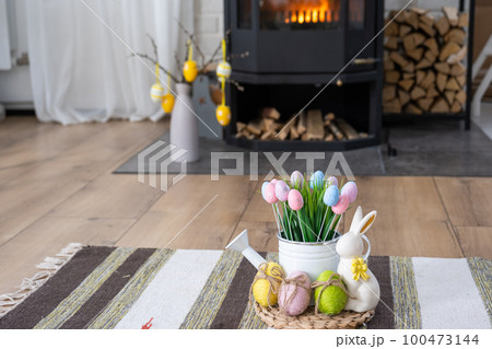 Easter decor near fireplace stove with fire and firewood. Cozy home hearth in interior with potted plans, colorful easter eggs, easter bunny and bouquet. Spring in a country house 100473144