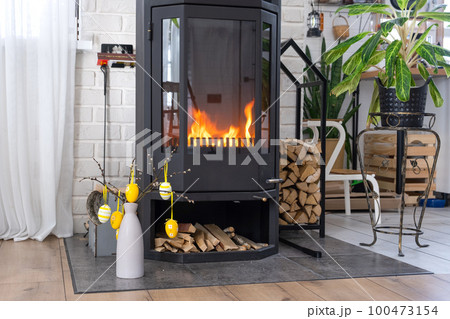 Easter decor near fireplace stove with fire and firewood. Cozy home hearth in interior with potted plans, colorful easter eggs, easter bunny and bouquet. Spring in a country house Easter decor near fireplace stove with fire and firewood. Cozy home hearth in interior with potted plans, colorful easter eggs, easter bunny and bouquet. Spring in a country house 100473154