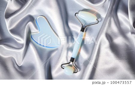 Blue jade face roller and Gua Sha scraping for beauty facial massage therapy on silver textured silk background. Blue jade face roller and Gua Sha scraping for beauty facial massage therapy on silver textured silk background. 100473557