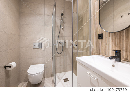 Small stylish design bathroom with stylish mirror, white ceramic sink, toilet and shower, finished with beige tiles. Bathroom concept in compact houses or hotels Small stylish design bathroom with stylish mirror, white ceramic sink, toilet and shower, finished with beige tiles. Bathroom concept in compact houses or hotels 100473759